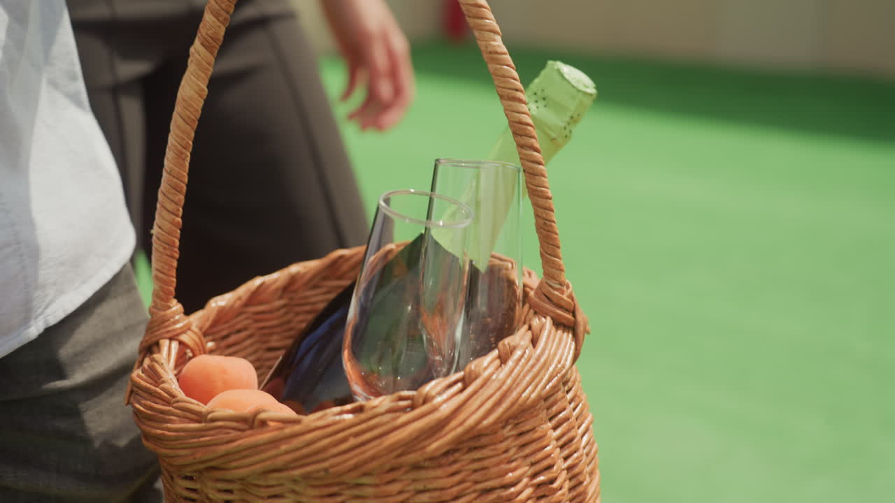 Detailní záběr proutěného košíku s lahvemi vajec drženého u těla na zeleném trávníku, pár v neformálním oblečení připravuje piknik, skleněná láhev a vejce uhnízděné v proutěném košíku, hřejivé sluneční světlo na džínách, pohodová letní atmosféra