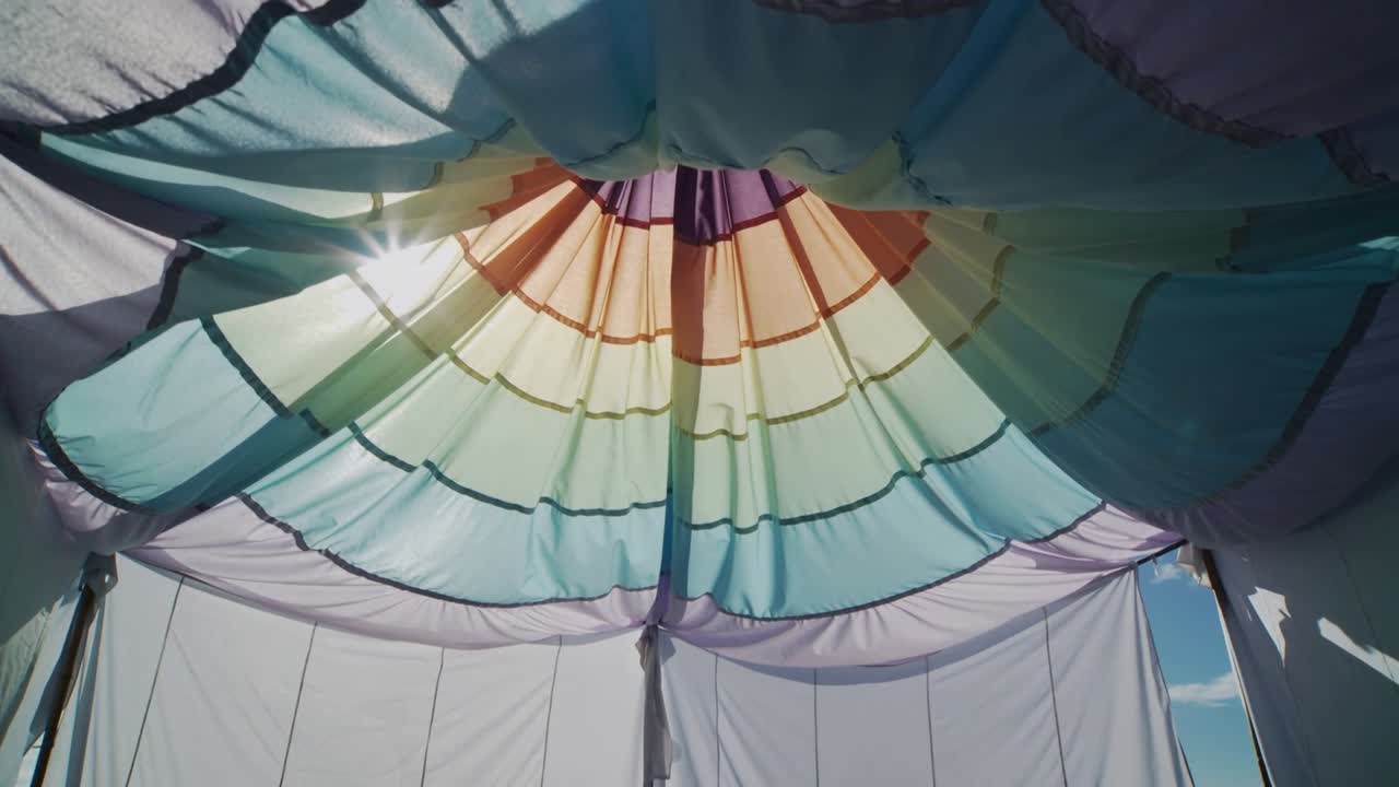 Cahaya matahari bergeser, kanopi pastel melingkar mengendap dan merata di tenda festival, menunjukkan tiang-tiangnya.