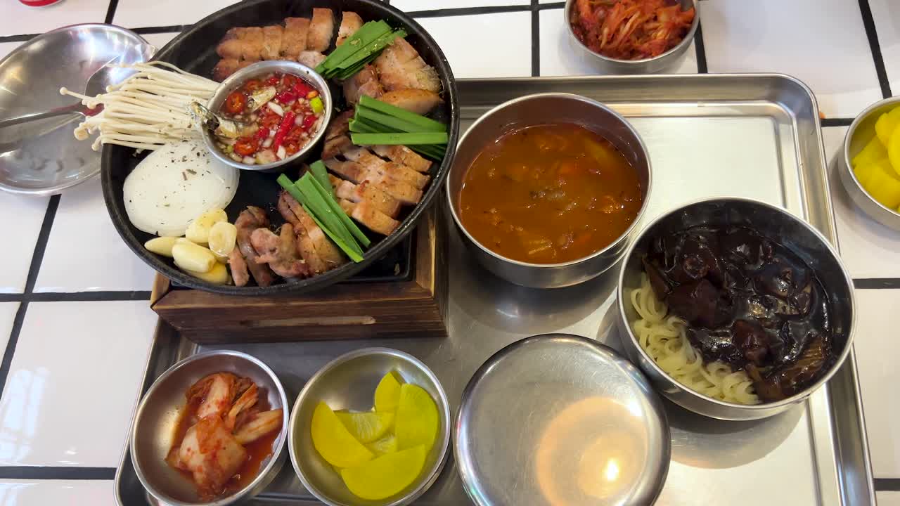 A hand organizes a vibrant Korean meal with grilled pork, jajangmyeon, kimchi, rice, and side dishes on a white-tiled table under bright lighting