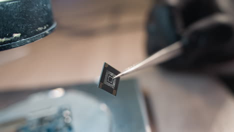 a close-up view of a gloved hand holding a microchip with tweezers, the background features a microscope and lab equipment