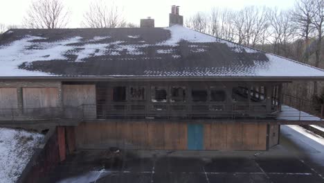 aerial drone footage dollying back from a broken window to reveal an abandoned lodge.  boarded windows, vandalization, winter.