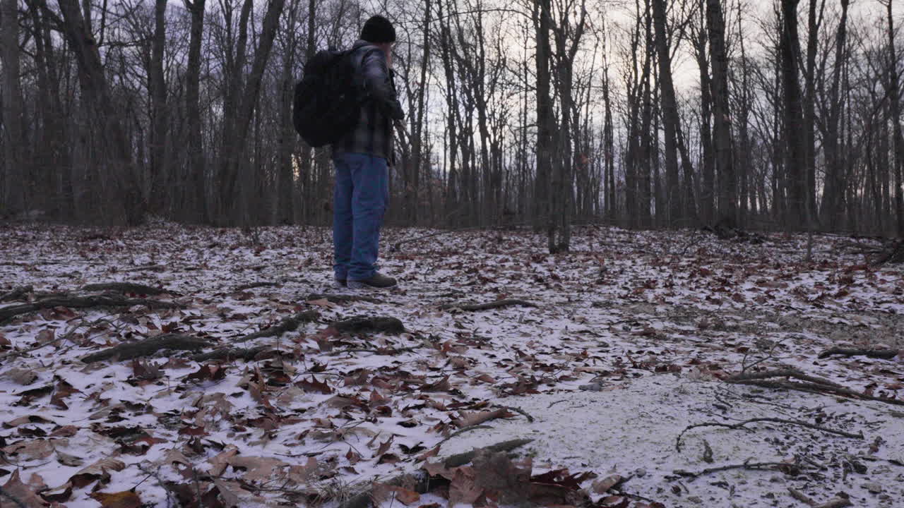 un tiro verso l'alto dal pavimento della foresta coperto di foglie asciutte e neve appena caduta a un escursionista in camicia di flanella e zaino che raccoglie accensione per un fuoco di campo