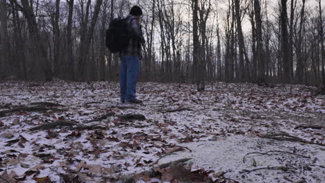 a tilt up shot from the forest floor covered with dried leaves and fresh fallen snow to a hiker in a flannel shirt and backpack collecting kindling for a campfire