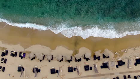 Top-down-aerial-rising-shot-of-azure-waves-crashing-in-towards-golden-beach-lined-with-sunbeds