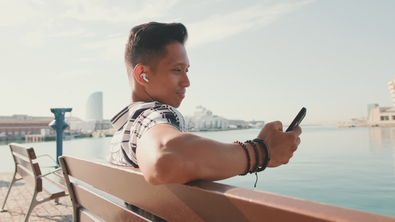 Man sitting on bench using phone near water