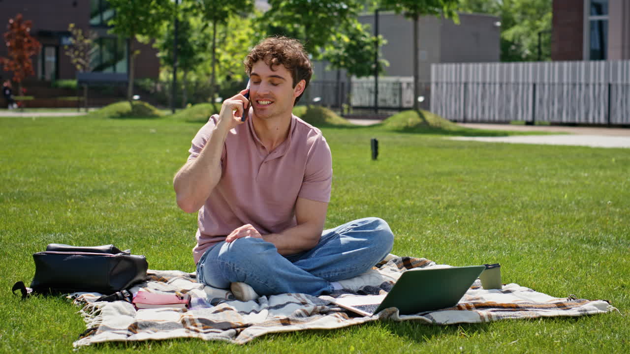 Cheerful freelancer calling cellphone sitting lawn with laptop. Smiling man