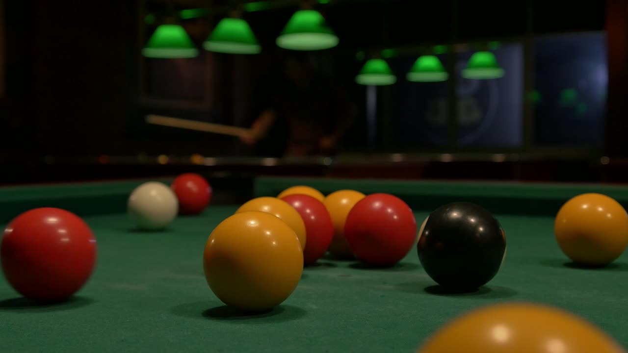 Close up, breaking a pool ball rack while people play on pool tables in the background with lights