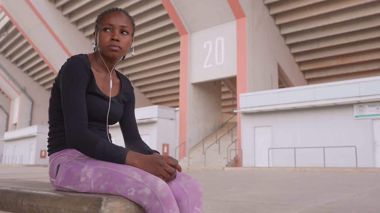 Female traveler resting. Individual waits on bench near stadium. Candid view of woman seated at stadium entrance. Side profile of woman with earphones on concrete bench in afternoon lighting