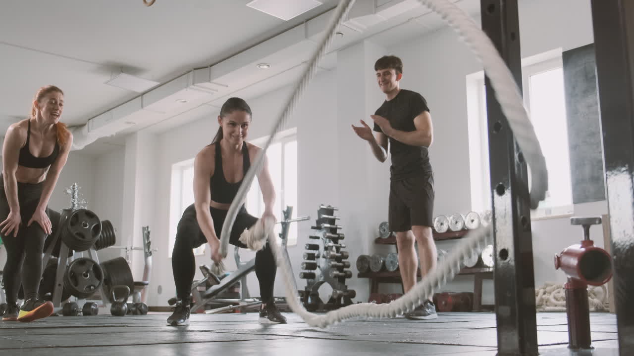 атлетически сложенная женщина работает с канатами, пока ее поддерживают товарищи по спортзалу