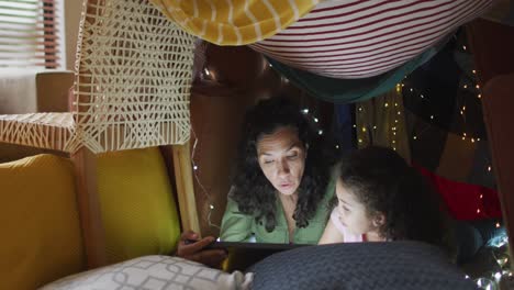 mixed race mother and daughter using tablet in blanket tent