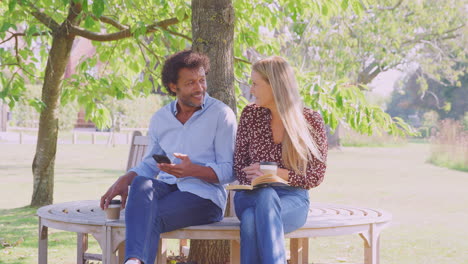 mature couple relaxing sitting on park bench under tree reading book and checking mobile phone