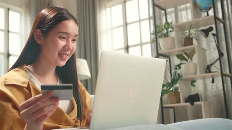 asian woman sitting on couch in living room holding credit card and celebrating while using laptop computer shopping online