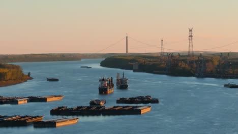 Toma-Aérea-De-Cerca-De-Barcos-Anclados-En-Un-Gran-Río-Azul,-Puente-Suspendido-En-El-Fondo,-Día-Claro-Y-Soleado,-4k50fps