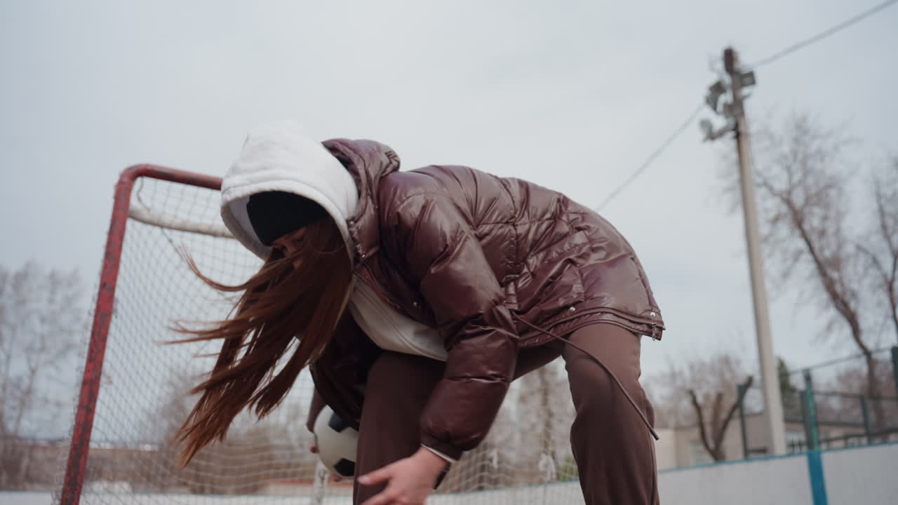 Adolescente caucásico recoge un balón de fútbol cerca de la portería agachándose rápidamente y con manos firmes, expresión concentrada y chaqueta de varias capas, luz fría y nublada, pista urbana y determinación por recuperar el juego