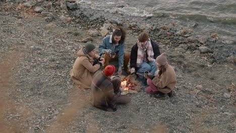 Group-Of-Teenage-Friends-Sitting-Around-The-Bonfire-While-They-Serving-Tea-In-Cups-With-A-Thermos-On-The-Seashore-1