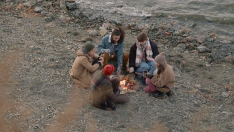 group of teenage friends sitting around the bonfire while they serving tea in cups with a thermos on the seashore 1