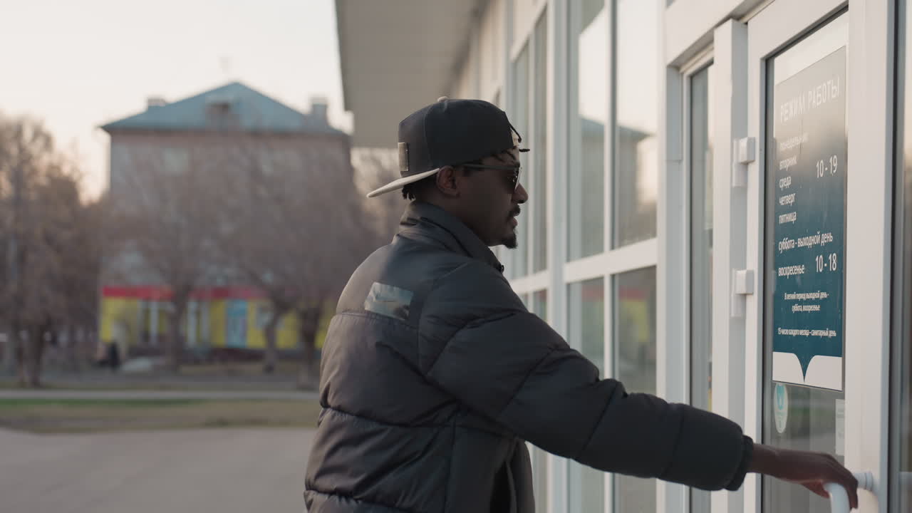 Close up side view of man in dark jacket and cap walking toward building entrance in urban setting during winter evening with blurred background of bare trees, parked cars, and residential houses