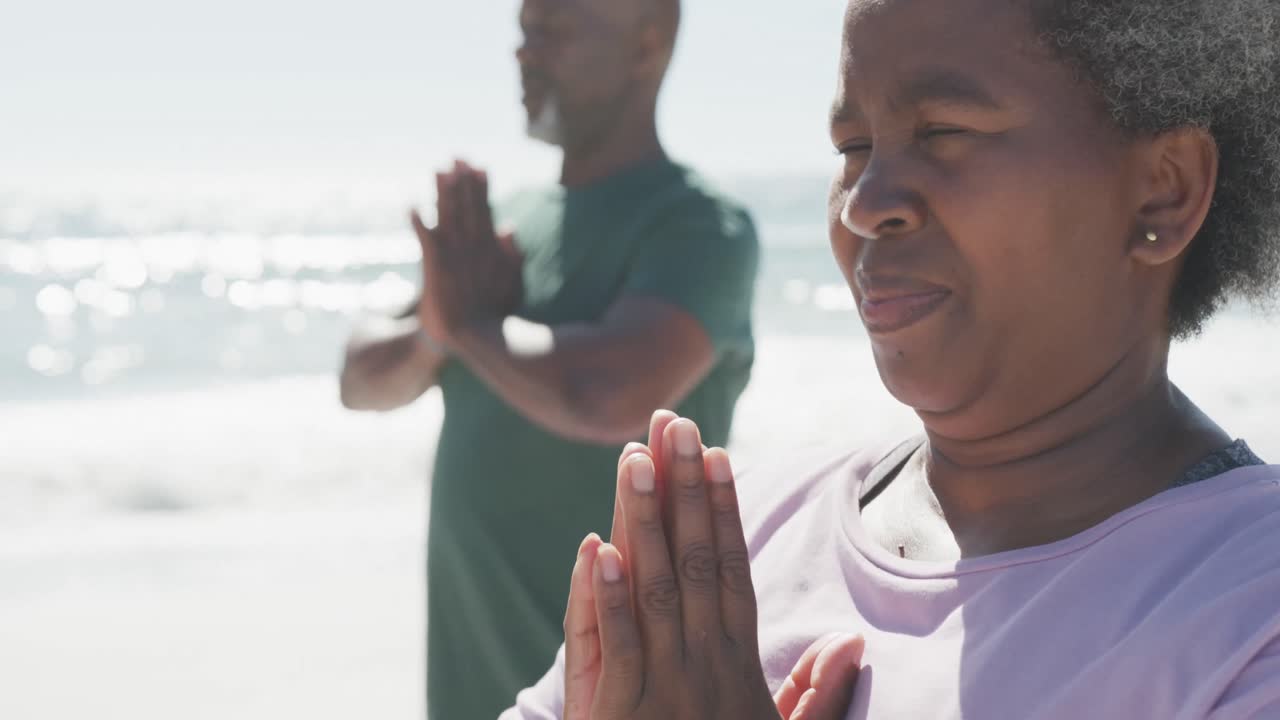 Happy senior african american couple doing yoga and meditating at beach, in slow motion