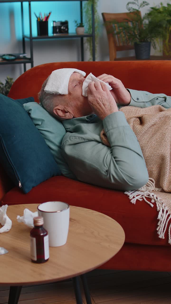 Anciano enfermo que sufre de alergia al resfriado, acostado en el sofá de casa, estornuda y se limpia los mocos con una servilleta