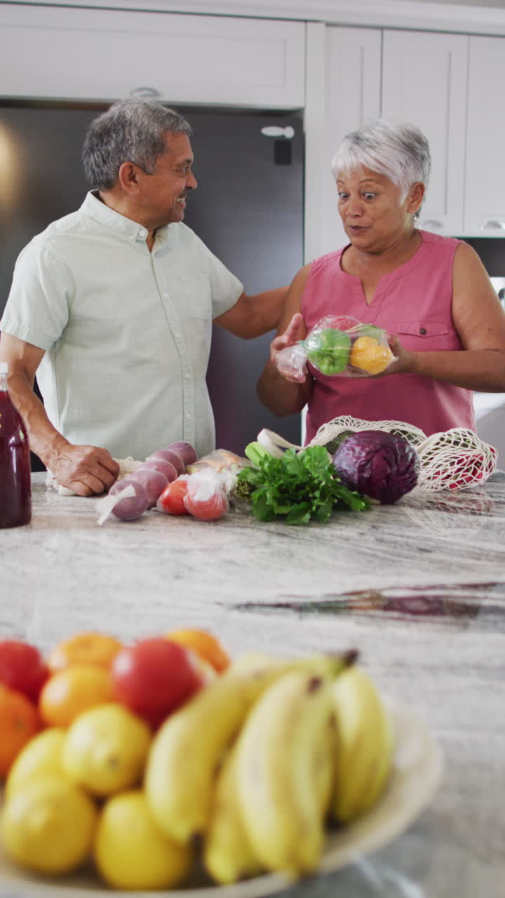 Vertical video of happy senior biracial couple talking and holding vegetables