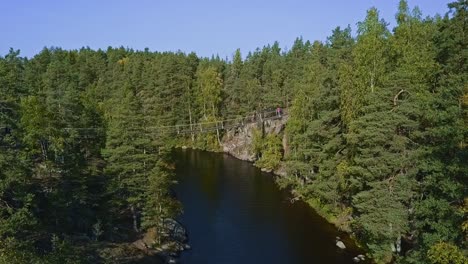 Floating-bridge-on-ropes-over-a-lake-or-a-stream-in-a-forest,-summer,-aerial-revealing-shot