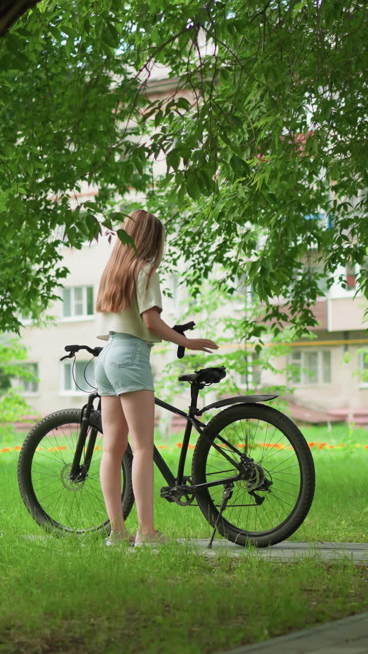 백인 여성이 포장된 길에 주차된 검은색 자전거에 접근하고, 다른 장갑을 착용하면서 자전거 좌석에 한 장갑을 놓고, 배경에는 멀리서 놀고 있는 어린이, 높은 나무 및 주거 건물이 있습니다.
