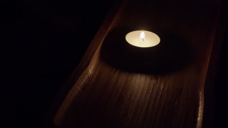 single tea candle burns in rich brown wooden tray in dark black room