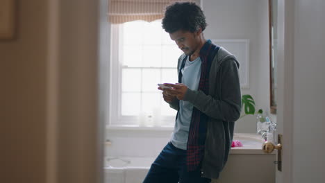 young mixed race man using smartphone in bathroom texting browsing online checking social media messages in morning before work