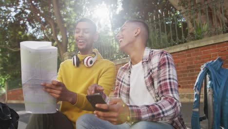 dos felices amigos de raza mixta sentados, usando un teléfono inteligente y un mapa