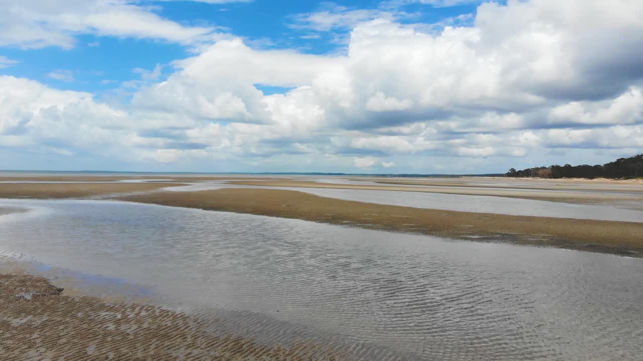 bajo, vuele sobre pequeños charcos y venas de agua en una playa de marea baja durante un día perfectamente soleado con nubes blancas, grandes y esponjosas flotando sobre la cabeza dando bonitos reflejos