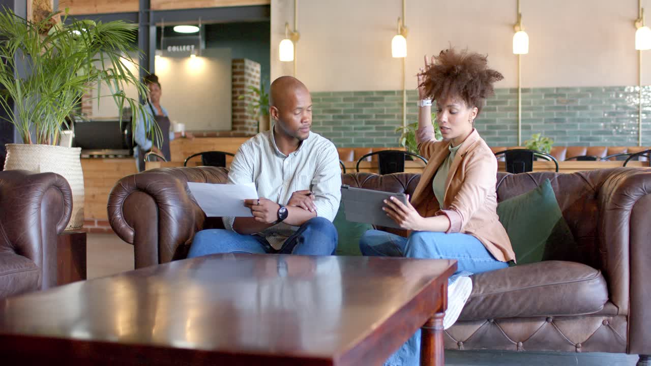 Afroamerikanske kolleger gjennomgår nettbrett og papirer på en brun kafésofa og snur seg mot forbipasserende personale.