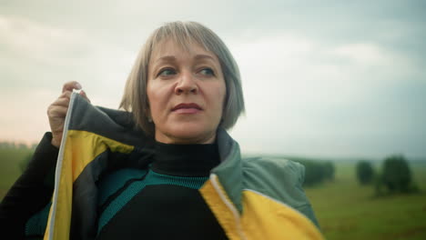 close-up of middle-aged woman in green and black suit unzipping yellow jacket while looking into the distance in vast open field under cloudy skies, with green trees visible in the distance