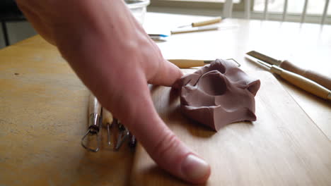 an artist carving and cutting brown modeling clay with a metal tool before sculpting and shaping the clay with his fingers slide right