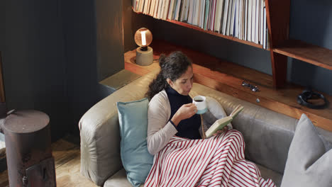 biracial woman lying on sofa under blanket reading book and drinking coffee at home, slow motion