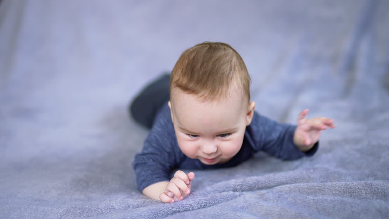 Little toddler in blue shirt lies on grey soft plaid. Cute baby boy moves actively to look around. Close up. Blurred backdrop.