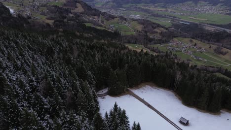 Aerial-rise-up-shot-of-single-hut-in-winter-pine-forest-with-view-over-vorarlberg-austria