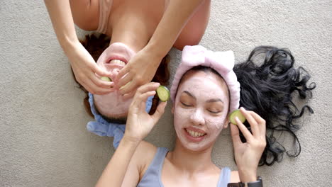 two young biracial women with facial masks hold cucumber slices