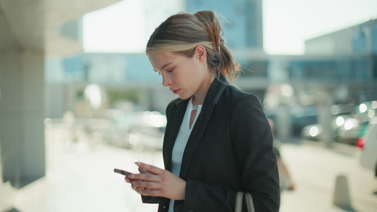 Contemplative woman in black suit operating phone thoughtfully outdoors with focus on screen, soft blur urban background featuring parked cars and daylight reflections