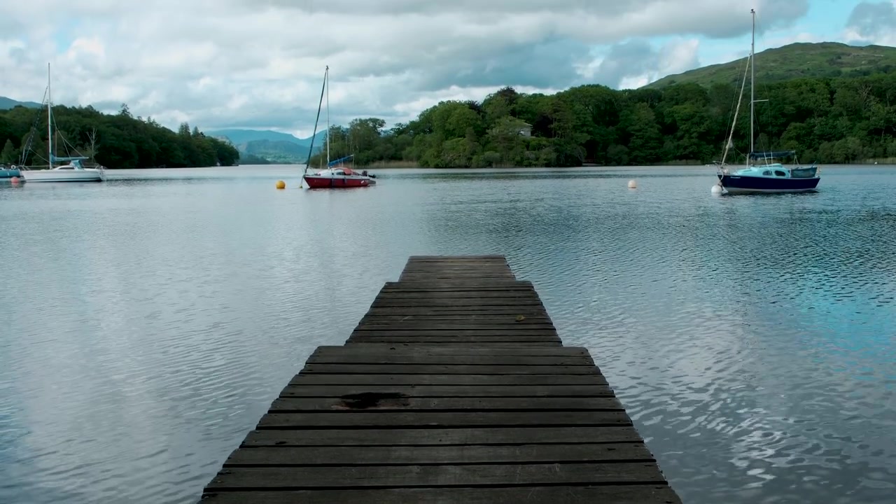 Ripresa panoramica verso l'alto da un pontile di legno attraverso Coniston Water nel Lake District, che rivela barche a vela ormeggiate, riflessi calmi, boschi circostanti e fells distanti