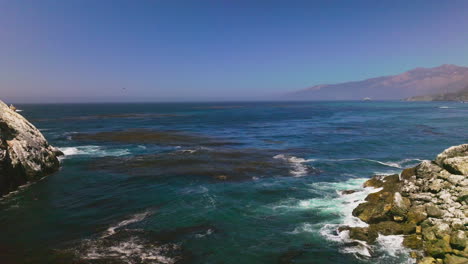 flying low above the blue water of the pacific ocean. algae floating on the water surface. mountains of california at hazy backdrop.