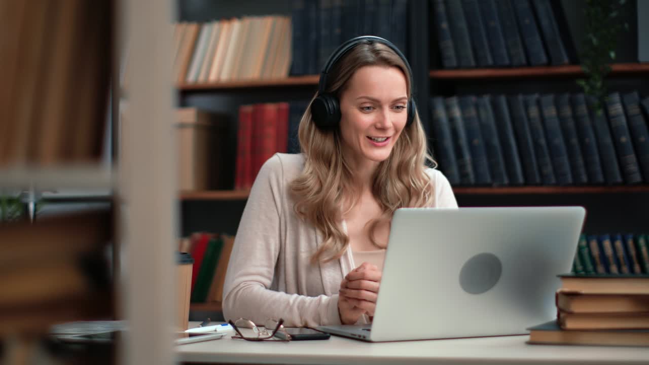 mujer sonriente con auriculares hablando en línea llamada de video portátil videochat consultoría trabajando en la biblioteca