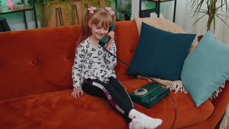 Smiling-young-children-girl-making-conversation-call-on-retro-phone-with-friends-on-couch-at-home