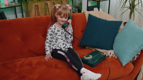 smiling young children girl making conversation call on retro phone with friends on couch at home