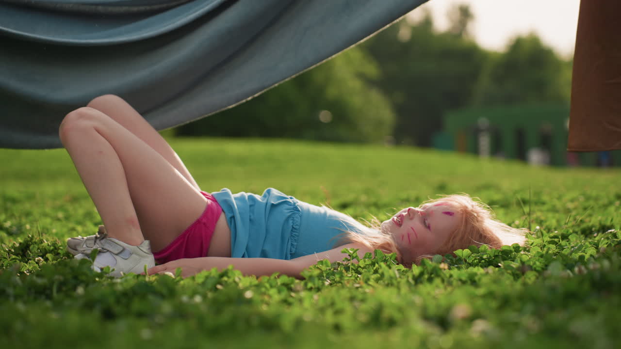 Young girl with face paint lying on grass while blanket sways in front, sunny park setting, smiling and enjoying playful moment, green background and soft light capturing carefree summer mood