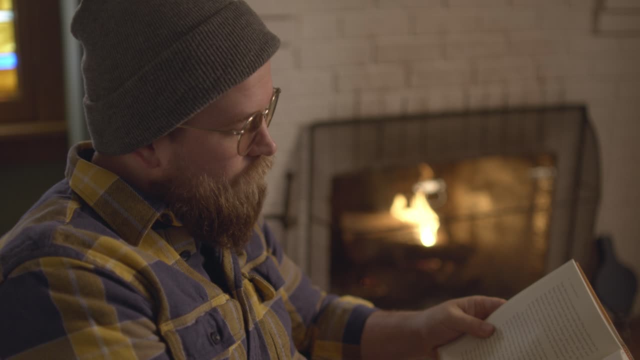 Close up of a bearded man sitting by a fire, flipping through book pages, and reading, a cozy fall living room setting, reading a book. Shot in 60 frames for slow motion