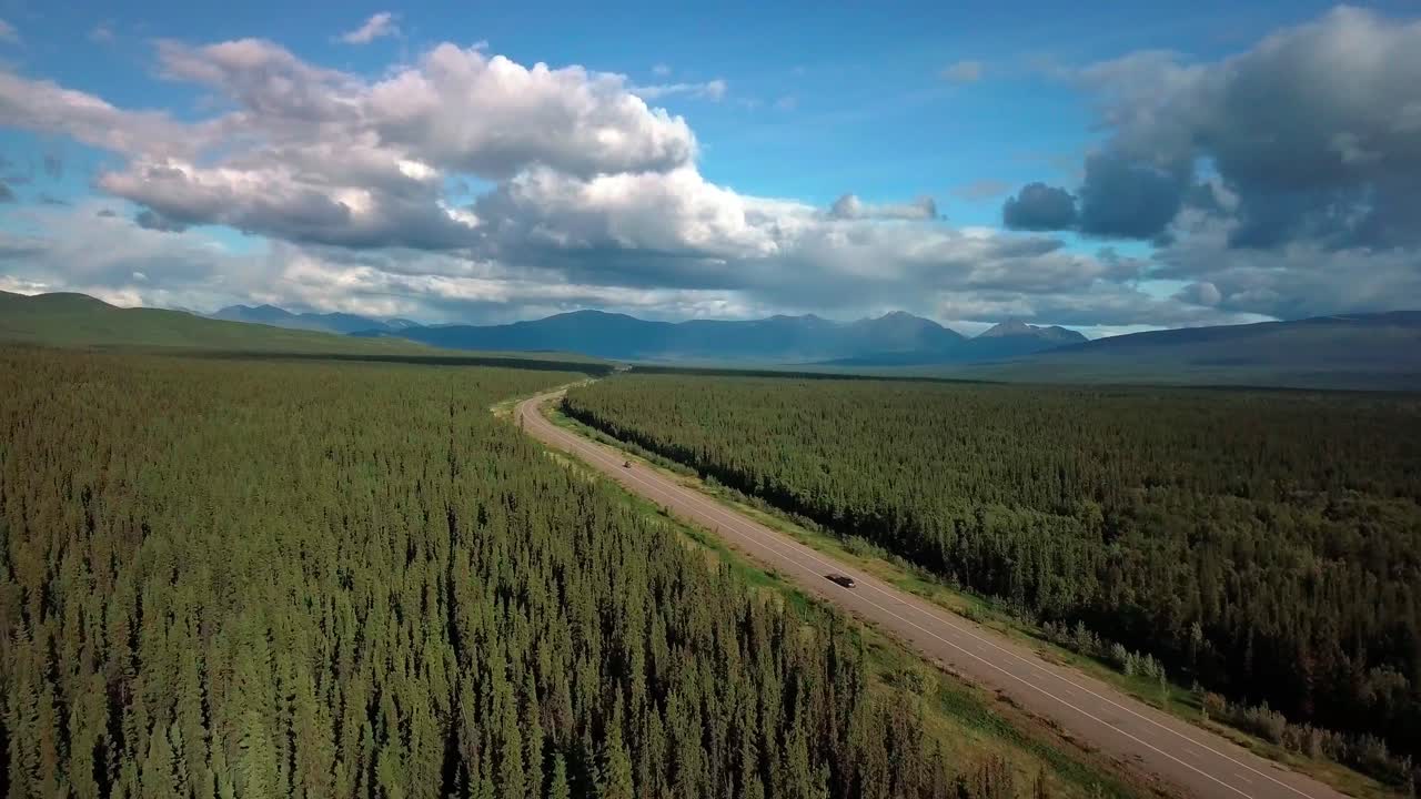 Epic landscape view of cars driving on narrow Alaska highway by evergreen trees in thick dense green forest and mountain range in background on blue sunny sky day, Yukon, Canada, above aerial approach