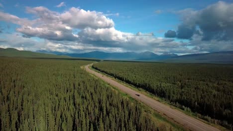 epic landscape view of cars driving on narrow alaska highway by evergreen trees in thick dense green forest and mountain range in background on blue sunny sky day, yukon, canada, above aerial approach