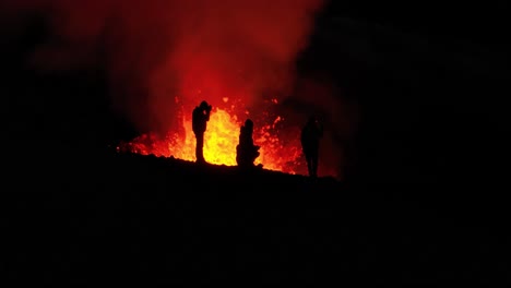 black silhouette of people on mountain ridge with exploding magma crater in background