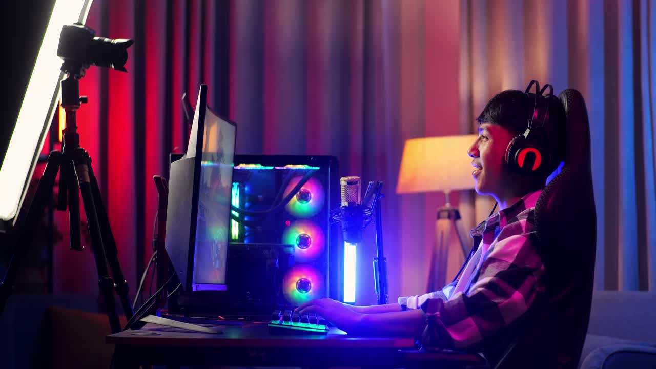 vista lateral de un niño asiático jugando a un juego en una computadora personal, agitando la mano hablando con los participantes en el micrófono. videojuego de transmisión en vivo, escritorio iluminado por luz de tira led rgb