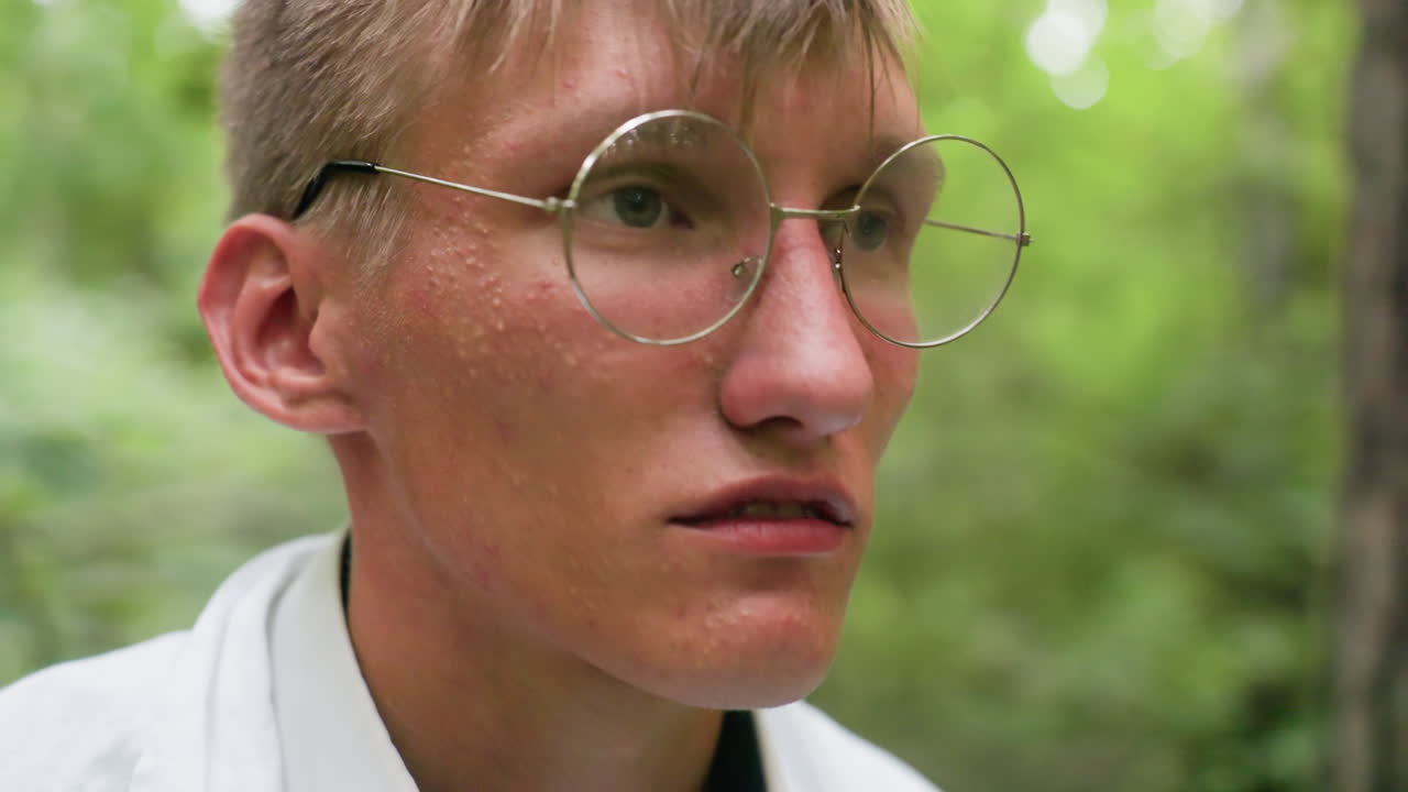 Portrait view botany student in white shirt putting on glasses in forest with tired expression surrounded by blurred trees, reflecting fatigue and stress during outdoor work in natural setting